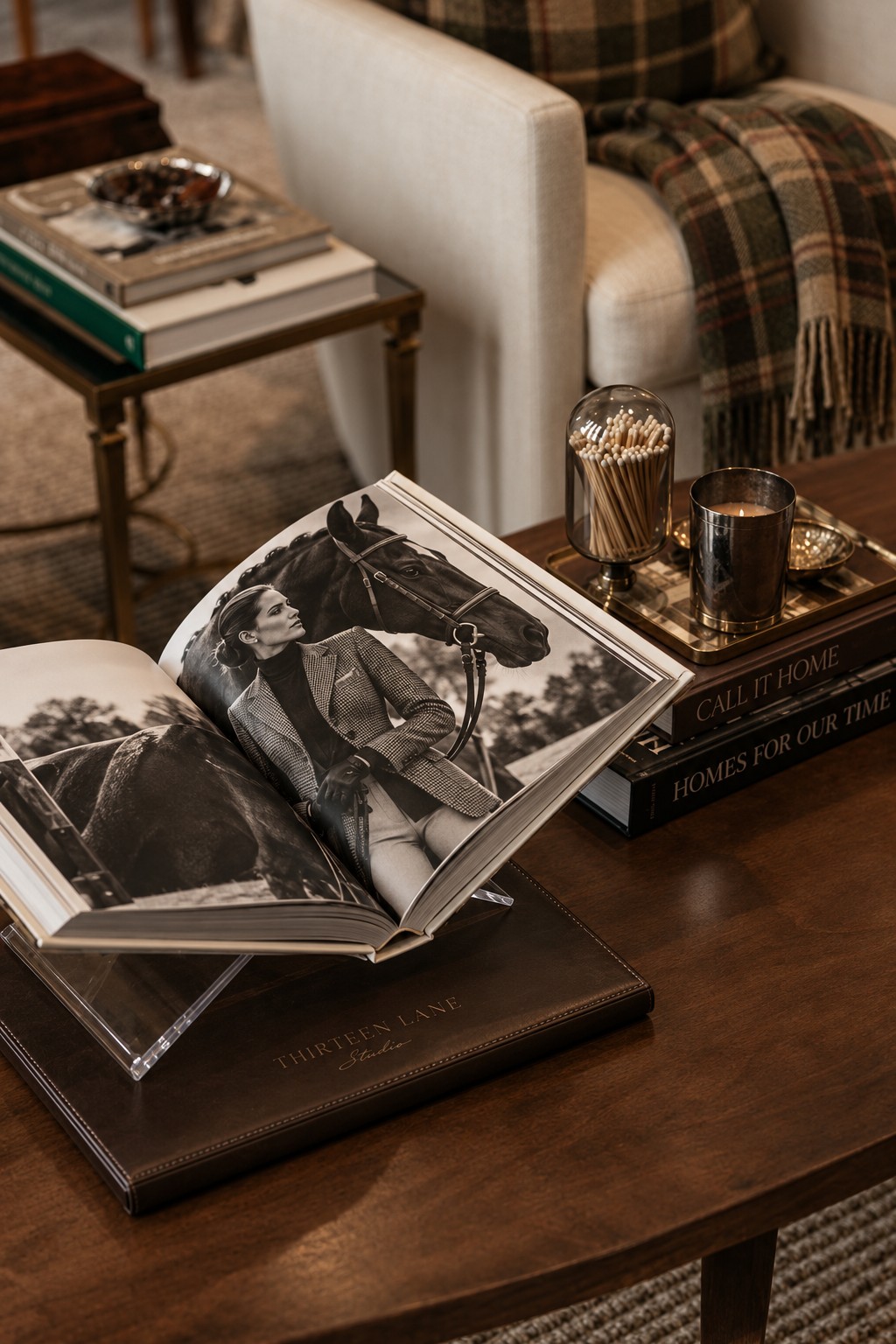 An equestrian editorial book on an espresso coffee table beside an ivory chair draped in olive and saddle-brown tartan cashmere